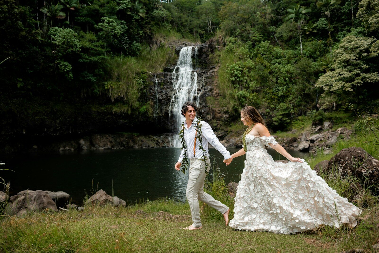 A man and a woman in a wedding dress holding hands near a waterfall